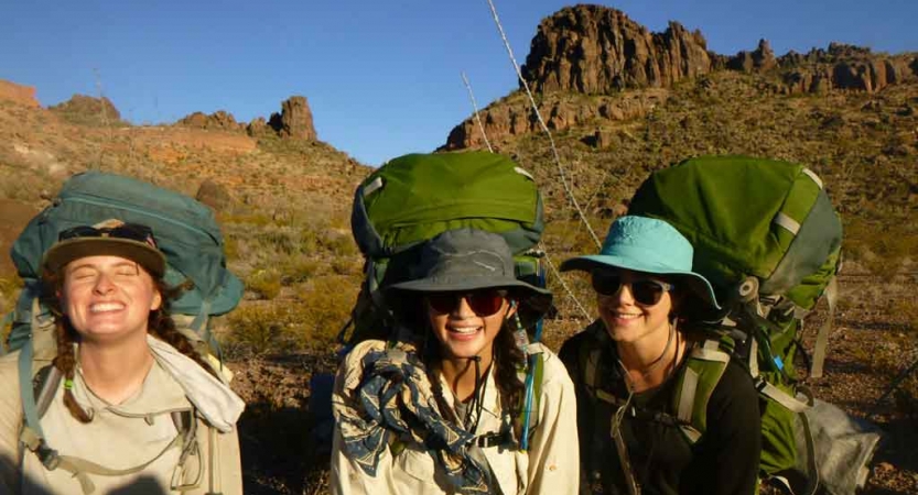 three gap year students wearing backpacks smile at the camera in front of a tall desert rock formation on an outward bound course
